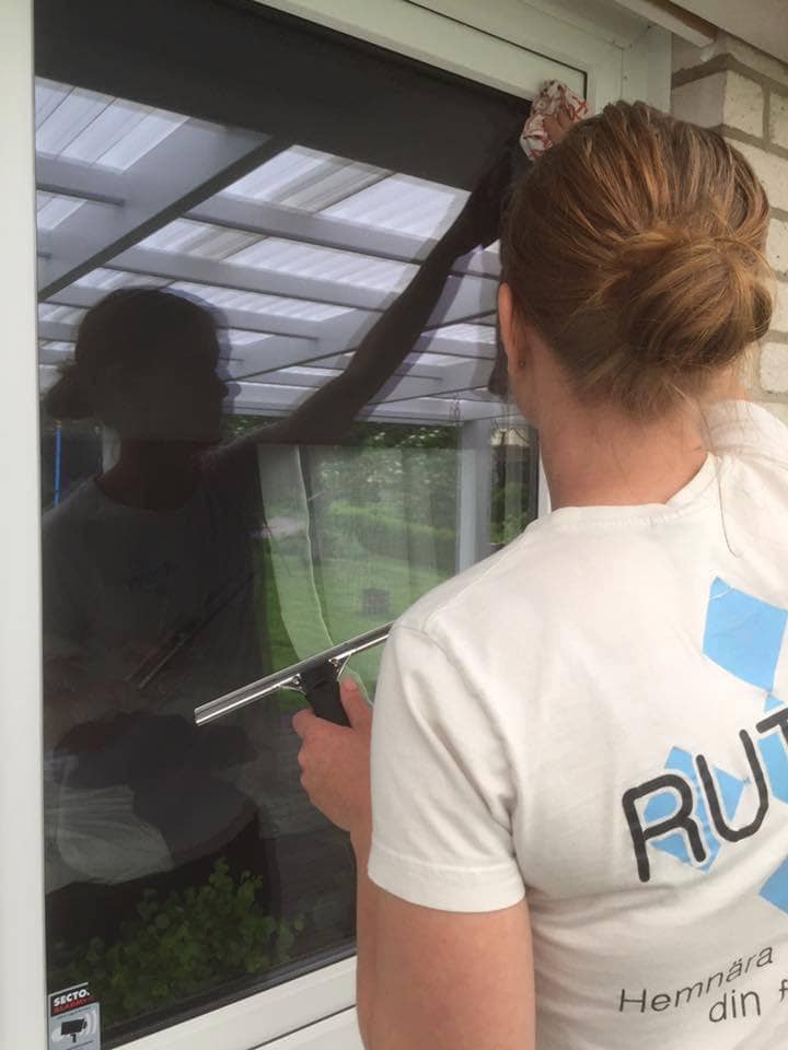 A hand wearing a green rubber glove is using a window squeegee to clean a glass window covered with soap suds.